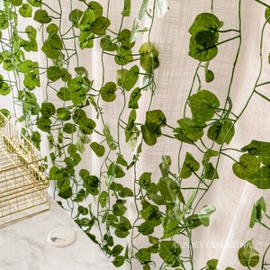 May include: A white sheer curtain with a green artificial ivy vine hanging in front of it. The ivy vine has many leaves and is cascading down the curtain.
