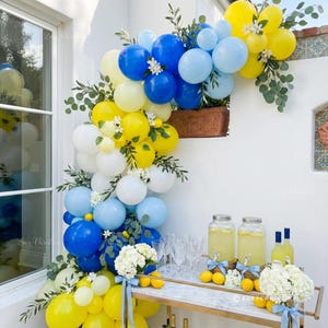 May include: A festive outdoor display featuring a balloon arch in shades of yellow, blue, and white, adorned with greenery and small white flowers. A bar cart with lemonade, lemons, and glassware completes the scene.