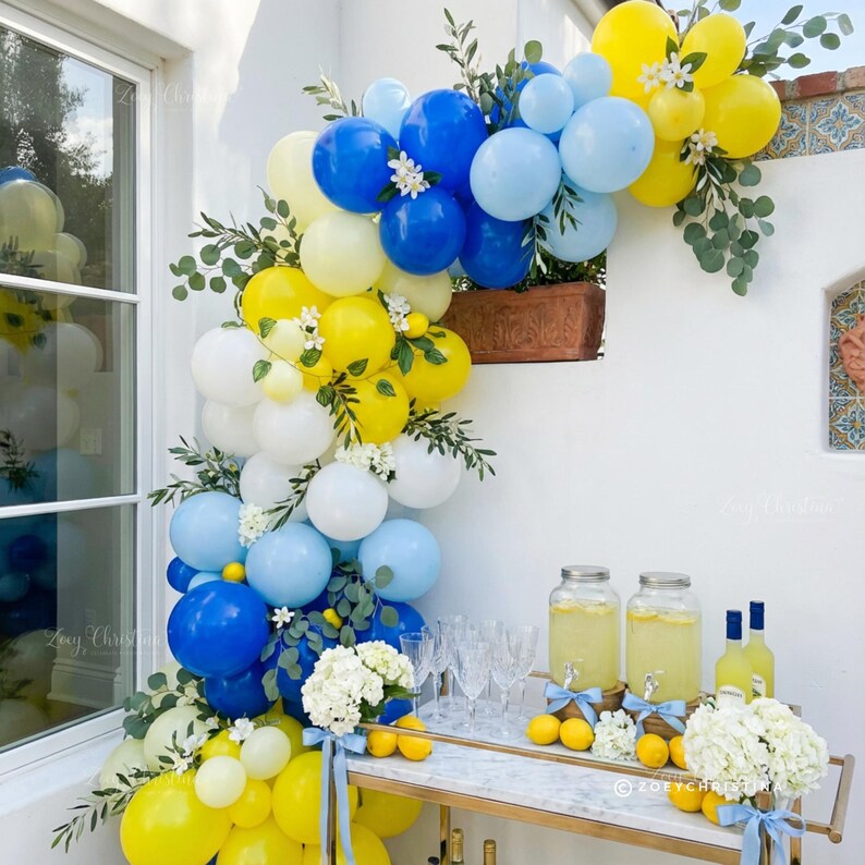 May include: A festive outdoor display featuring a balloon arch in shades of yellow, blue, and white, adorned with greenery and small white flowers. A bar cart with lemonade, lemons, and glassware completes the scene.