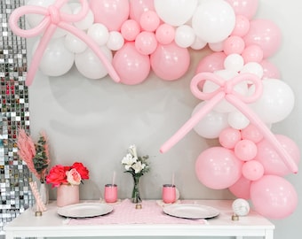Puede incluir: Arco de globos rosa y blanco con un lazo hecho de globos. El arco está decorado con globos rosa y blanco. El arco está instalado frente a una pared blanca. Hay una mesa con manteles individuales rosa y blanco frente al arco.
