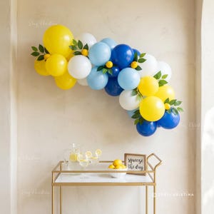 May include: A balloon arch in yellow, white, and blue, adorned with lemon and leaf accents, hangs above a gold bar cart. The cart holds a pitcher of lemonade, glasses, and a sign that reads "squeeze the day."