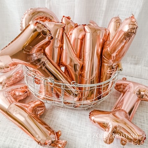 May include: A group of rose gold foil balloons in the shape of the letters "ONE" are arranged in a wire basket. The balloons are shiny and reflect light.