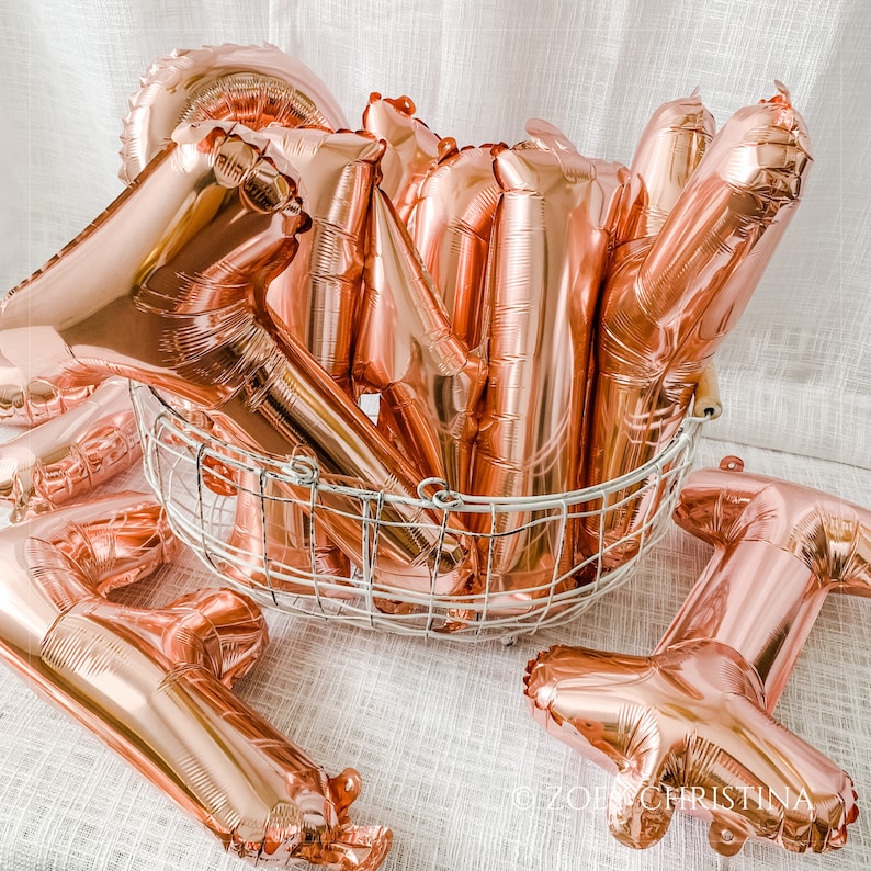 May include: A group of rose gold foil balloons in the shape of the letters "ONE" are arranged in a wire basket. The balloons are shiny and reflect light.
