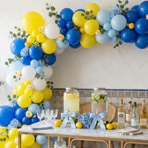 May include: A festive party scene with a balloon arch in yellow, blue, and white. The table is set with drinks, lemons, and flowers. Glassware and bottles are arranged on a marble-topped table, creating a celebratory atmosphere.