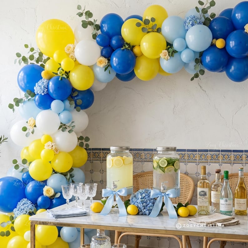 May include: A festive party scene with a balloon arch in yellow, blue, and white. The table is set with drinks, lemons, and flowers. Glassware and bottles are arranged on a marble-topped table, creating a celebratory atmosphere.
