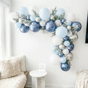 May include: A festive balloon arch in shades of white, light blue, and navy blue, accented with silver orbs and greenery. The arrangement is displayed against a white wall, creating a celebratory atmosphere. A small sign reads "Merry Christmas".
