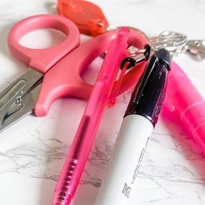 May include: A collection of pink office supplies, including scissors, a pen, and a marker. The scissors are a bright pink, and the pen is a translucent pink. The marker has a white body with a black tip. A red keychain is also visible.