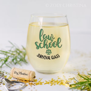 May include: A stemless wine glass filled with a light-colored liquid, featuring the text "law school" in green script, with a scale symbol and the words "survival glass" below. A cork and gold confetti are in the foreground.