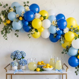 May include: A festive arrangement featuring a balloon arch in shades of blue, yellow, and white, adorned with greenery and small white flowers. A table displays blue hydrangeas, lemonade, and lemons, creating a cheerful, summery scene.