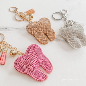 May include: Three rhinestone-covered tooth-shaped keychains in gold, silver, and pink. Each keychain has a metal clasp, chain, and tassel. The keychains are displayed on a white marble surface.