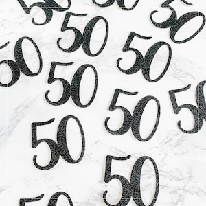 May include: A close-up of multiple black glitter-covered paper cutouts of the number 50. The cutouts are scattered on a white marble surface.