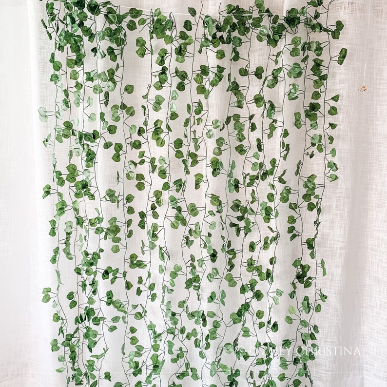 May include: A white curtain with a green garland of artificial leaves hanging from the top. The garland is made of plastic leaves and is attached to the curtain with a wire.