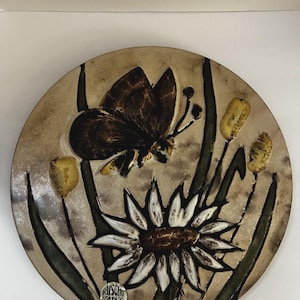 May include: A round ceramic wall plaque featuring a raised relief design of a dark brown butterfly, a large white daisy, and yellow flower buds on a beige background. The piece includes a small label with the text Ruscha Art echt Handarbeit.