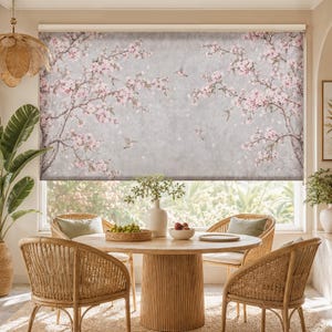 Puede incluir: Un estor enrollable con un diseño estampado de ramas de cerezo en flor de color rosa sobre un fondo gris claro texturizado, con pequeños pájaros representados en vuelo entre los motivos florales, cubriendo una ventana sobre un comedor.