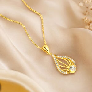 Puede incluir: Un collar de tono dorado con un colgante en forma de lágrima. El colgante tiene un centro hueco con líneas doradas radiantes y está adornado con pequeñas piedras claras y brillantes a lo largo del borde exterior y un grupo en la base.