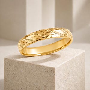 Könnte beinhalten: Ein polierter Ring aus Gelbgold mit einer strukturierten Oberfläche, die diagonale, sich wiederholende Muster aus feinen, erhabenen Perlenlinien und glatten, schrägen Rillen aufweist, welche den gesamten Umfang des runden Metallschmuckstücks umschließen.