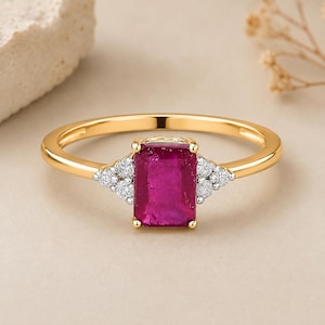 Puede incluir: Un anillo de oro con una piedra central de rubí rectangular de color rosa intenso en un engaste de cuatro garras. La banda está acentuada a ambos lados por un grupo de tres pequeños diamantes blancos redondos en engastes plateados.