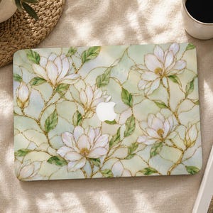 May include: A laptop skin featuring a stained glass design with white magnolia flowers, green leaves, and gold-coloured outlines. The pattern covers the entire surface, surrounding a central cutout for the Apple logo on the laptop lid.