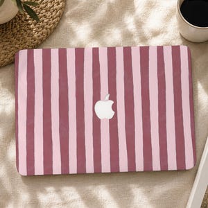 Peut inclure: Un autocollant pour ordinateur portable avec un motif à rayures verticales alternant des tons de vieux rose et de rose clair. Le design présente un aspect texturé peint à la main aux bords irréguliers. Un logo Apple blanc est visible au centre.