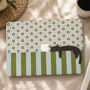 May include: A laptop skin featuring a cream background with a repeating dark blue floral pattern on the top half and vertical olive green and light blue stripes on the bottom half, with a black cat illustration resting near the Apple logo.