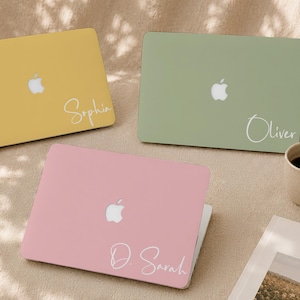 May include: Three laptop cases in yellow, sage green, and light pink, each featuring a white Apple logo and a custom name in white script font. The names displayed are Sophia on the yellow case, Oliver on the green case, and D. Sarah on the pink case.