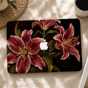 Pode incluir: Uma capa para portátil com uma ilustração em estilo de pintura a óleo de três grandes lírios cor-de-rosa vibrantes com manchas escuras e caules verdes sobre um fundo preto profundo. O logótipo branco da Apple é visível no centro.
