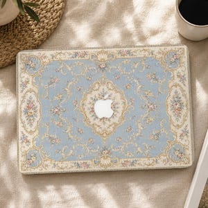 Peut inclure: Une coque rigide rectangulaire pour ordinateur portable avec un fond bleu clair orné d'un motif damassé floral crème et doré. Le design comprend de petits bouquets de fleurs roses et jaunes, avec une découpe centrale pour le logo Apple.