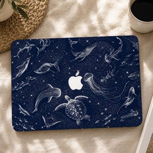 Peut inclure: Un autocollant pour ordinateur portable bleu foncé avec un motif en lignes blanches représentant diverses créatures marines, dont une tortue, une méduse, une baleine et des requins sur un fond étoilé, avec une découpe pour le logo Apple.