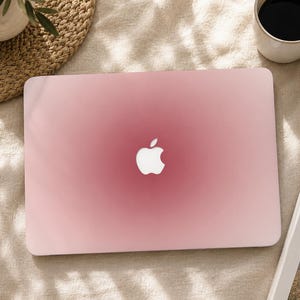 May include: A rectangular laptop with a smooth, matte finish featuring a soft pink gradient that transitions from a darker rose centre to a lighter blush perimeter. The iconic white Apple logo is centred on the lid of the closed device.