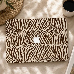 May include: A rectangular laptop cover featuring a repeating dark brown and cream zebra stripe pattern. The cover has a central cutout revealing the white Apple logo, which is positioned in the middle of the animal print design on the flat surface.