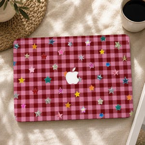 Peut inclure: Une coque d'ordinateur portable avec un motif à carreaux vichy rouge foncé et rose clair. La surface est ornée de nombreuses petites étoiles autocollantes multicolores dans des tons bleu, jaune, rose, argenté et vert, autour du logo Apple blanc.