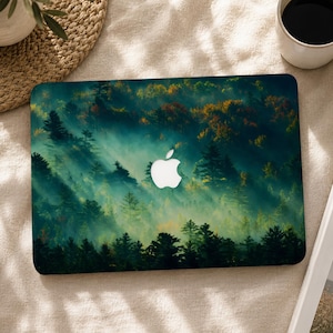 May include: A laptop skin featuring a dense forest landscape with tall pine trees shrouded in thick, ethereal morning mist. The scene displays deep green and golden autumn foliage, centered around a white Apple logo cutout on the laptop lid.