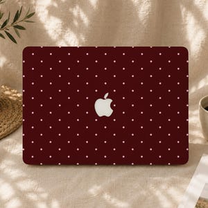 May include: A dark maroon laptop skin featuring a repeating pattern of small, light pink polka dots. The skin is applied to the lid of a laptop, with a central cutout revealing the white Apple logo. The surface has a smooth, matte finish.