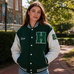 Pode incluir: Um casaco varsity verde-escuro com mangas brancas, apresentando uma letra H grande verde no peito, o nome Henry bordado em escrita branca no lado direito e o número 23 na manga esquerda, com acabamento em punhos canelados às riscas.