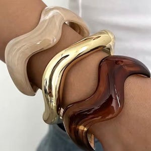 May include: A set of three wavy bangles worn together. One bangle is a glossy beige marbled resin, the second is a polished gold-tone metal, and the third is a deep brown translucent resin. Each bracelet features a distinct undulating shape.