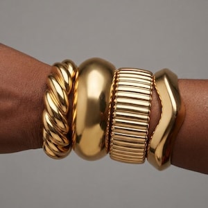 May include: A set of four gold-toned metal bracelets worn together. The stack includes a twisted rope-style bangle, a smooth polished wide cuff, a ribbed textured band, and a single wavy-edged bangle, all featuring a high-shine metallic finish.