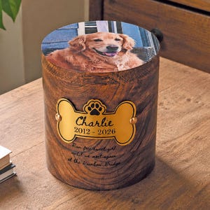 May include: A cylindrical wooden pet urn featuring a photo of a golden retriever on the lid. A gold-coloured bone-shaped plaque is attached to the front, engraved with the name Charlie, the years 2012-2026, and a paw print icon.