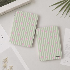 Könnte beinhalten: Zwei rechteckige Tablet-Hüllen mit einem vertikalen Streifenmuster in Hellgrün und Weiß, das mit kleinen rosa Herzen übersät ist. Eine der Hüllen trägt den Namen Joshua, der in einer schwarzen Schreibschrift unten rechts aufgedruckt ist.