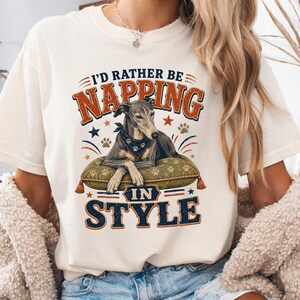 Pode incluir: Uma t-shirt de manga curta na cor marfim com o gráfico de um galgo usando uma bandana, descansando sobre uma almofada verde. O texto diz I'D RATHER BE NAPPING IN STYLE, acompanhado por pequenas ilustrações de estrelas e pegadas em vermelho, branco e azul.
