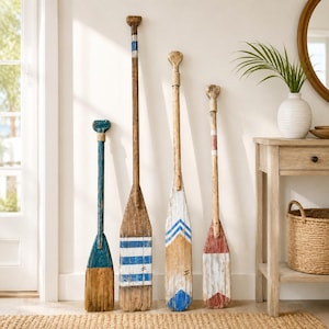Op de afbeelding: Vier decoratieve houten roeispanen die rechtop staan. De eerste is blauw en naturel hout, de tweede heeft blauwe en witte horizontale strepen, de derde heeft blauwe chevronpatronen op wit en de vierde toont een rood en wit chevronontwerp.