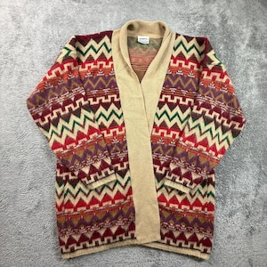 Puede incluir: Un cárdigan de manga larga con un patrón geométrico multicolor en tonos rojo, verde, morado y beige. La prenda tiene un cuello chal de color beige sólido y una abertura frontal. Una pequeña etiqueta blanca de marca es visible en el cuello interior.