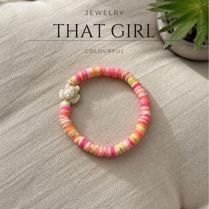 Puede incluir: Una pulsera circular hecha de pequeñas cuentas de arcilla planas y multicolores en tonos rosa, naranja, amarillo y blanco. La pulsera presenta un dije de tortuga tridimensional en color blanco hueso. El texto JEWELRY THAT GIRL COLOURFUL es visible.