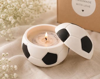 May include: A soccer ball shaped candle container with a lid, featuring a classic white and black pentagon pattern. The candle is lit, showing a single wick centered in white wax. A brown cardboard box with a sticker reading HANDMADE WITH LOVE is visible.
