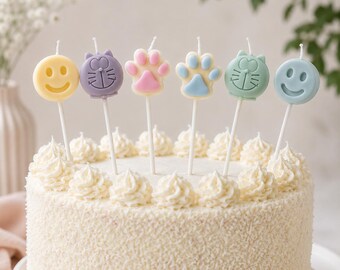 May include: A round white cake topped with six decorative candles on sticks. The candles feature a yellow smiley face, a purple cat face, a pink and cream paw print, a blue and cream paw print, a green cat face, and a blue smiley face.