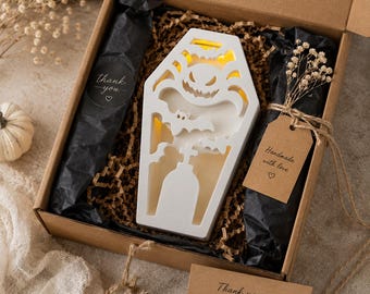 May include: A white coffin-shaped light box featuring cutouts of a spider, bats, and a tombstone, nestled in a cardboard gift box with black tissue paper, brown crinkle paper, and a tag labeled Handmade with love and a thank you sticker.