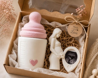 May include: A gift box containing a white baby bottle with a pink nipple and heart detail, a white figurine of a pregnant person, and a small oval frame holding an ultrasound image. A round tag attached with twine reads Made with Love.