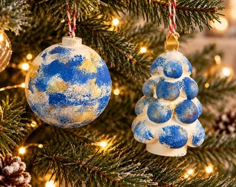 May include: Two Christmas tree ornaments hanging from red and white string. One is a spherical ball and the other is a tiered tree shape. Both ornaments feature a white base with a textured, mottled pattern of blue and gold paint throughout.