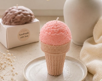 May include: A pink ice cream cone shaped candle with a textured scoop and a waffle cone base featuring the embossed text ICE CONE CECREAM. The candle sits on a small round plate next to a white box with a handmade with love label and a brown candle.