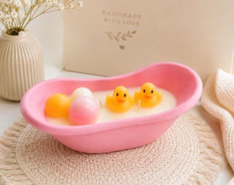 May include: A pink bathtub-shaped candle filled with white wax, featuring two yellow rubber duck-shaped wax embeds and two round wax spheres in orange and pink. The candle has two visible wicks and is presented in a decorative container.