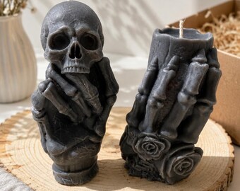 May include: Two dark gray wax candles shaped like skeletal forms. One candle depicts a human skull resting on a skeletal hand, while the other candle features a pair of skeletal hands clasped together with sculpted roses at the base.
